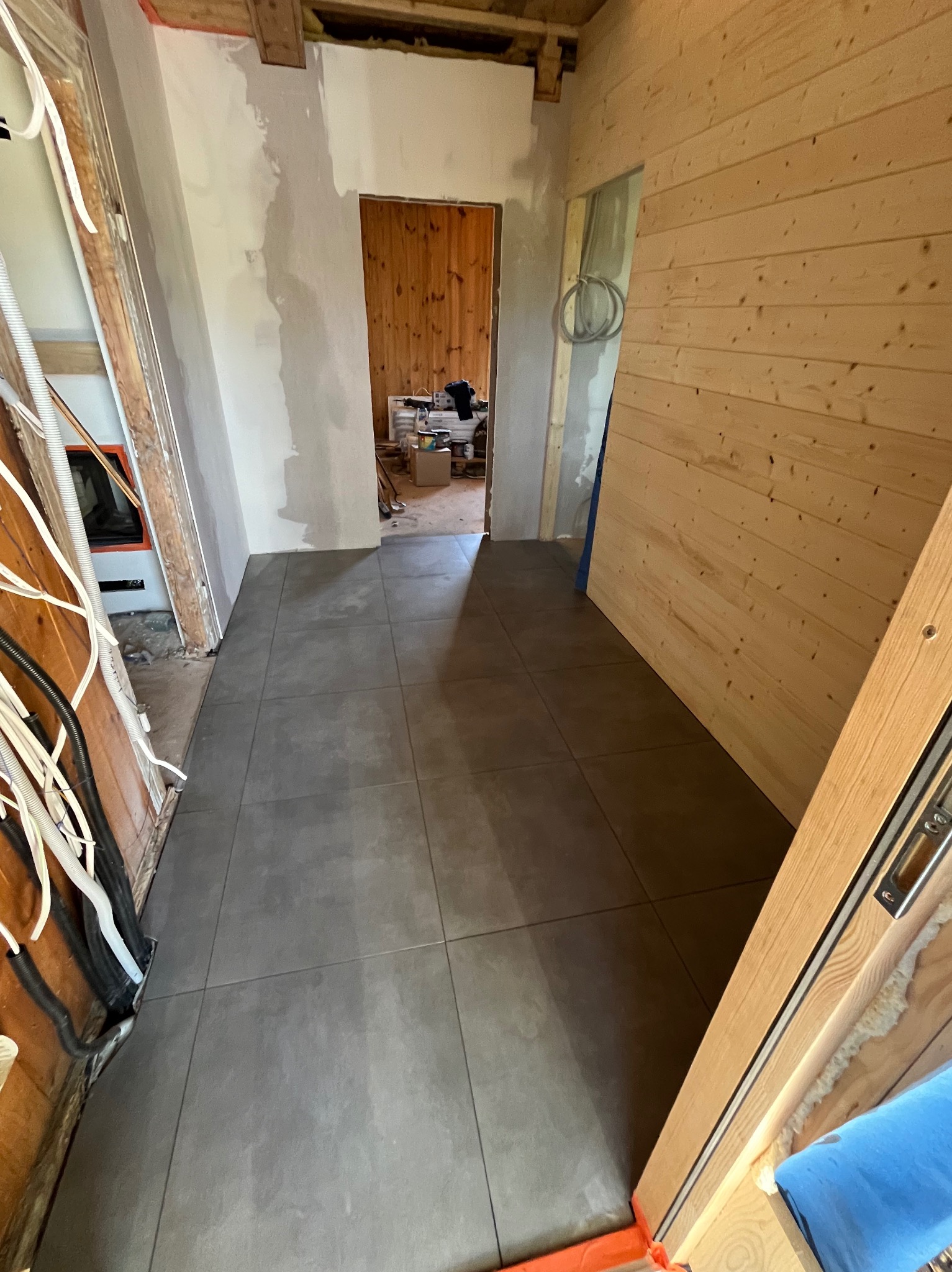 Wnętrze w trakcie remontu: szare płytki podłogowe, surowe drewniane ściany, widoczne instalacje elektryczne i przygotowane ściany do malowania.