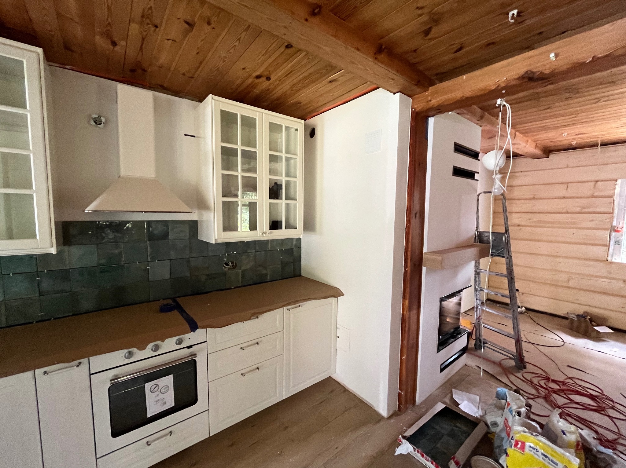 Wnętrze domu w trakcie remontu: zabudowa kuchenna z białymi szafkami i zieloną glazurą, kominek w trakcie budowy, drewniany sufit z widocznymi belkami, drabina i przewody elektryczne na podłodze.