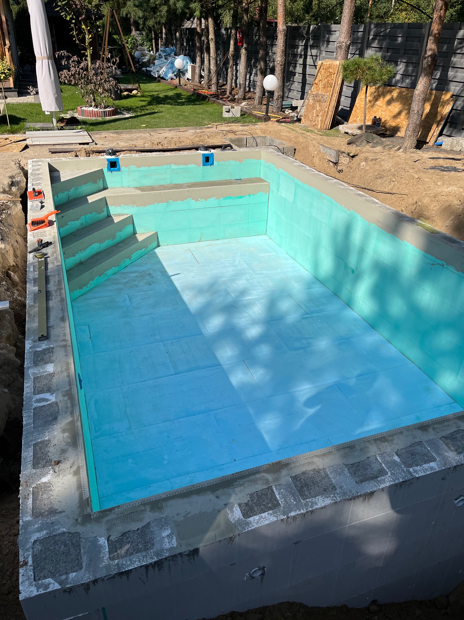 Wykopany dół pod basen ogrodowy z betonową konstrukcją i wewnętrzną izolacją z niebieskich płyt styropianowych, widoczne schody z betonu, narzędzia murarskie i poziomica.