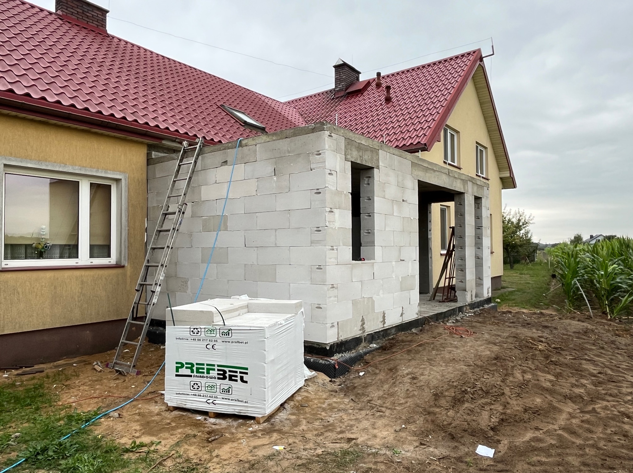 Budowa domu jednorodzinnego w trakcie realizacji, widoczne białe bloczki betonu komórkowego, drabina oparta o ścianę, paleta bloczków marki Prefbet, czerwony dach budynku w tle.
