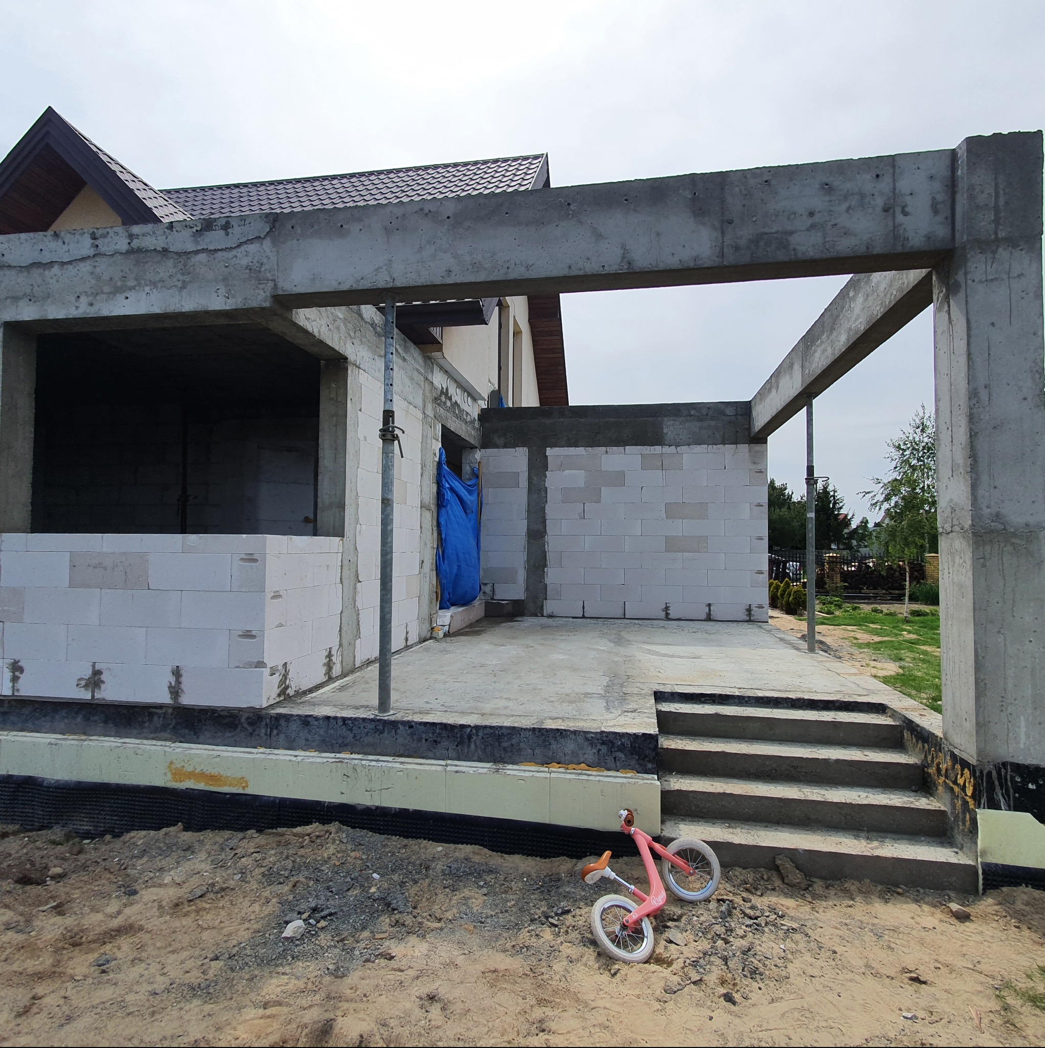 Surowa konstrukcja tarasu z betonu i bloczków, widoczne zbrojenia i stopnie, na ziemi leży różowy rowerek biegowy.