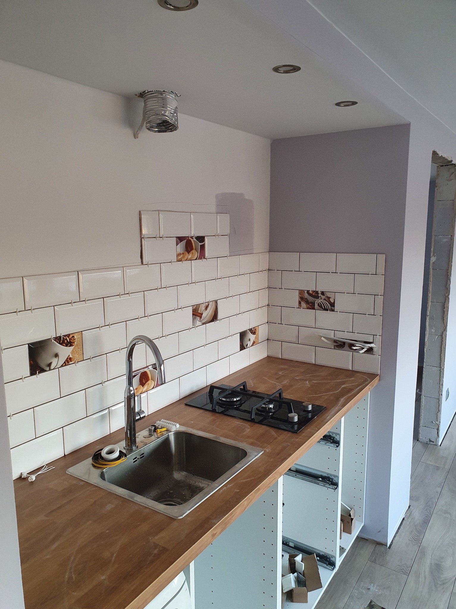 Montaż płytek ściennych z motywem kawy nad blatem kuchennym z zamontowanym zlewem i płytą gazową, widoczne listwy dystansowe między płytkami, instalacja wentylacyjna w suficie, surowy stan...