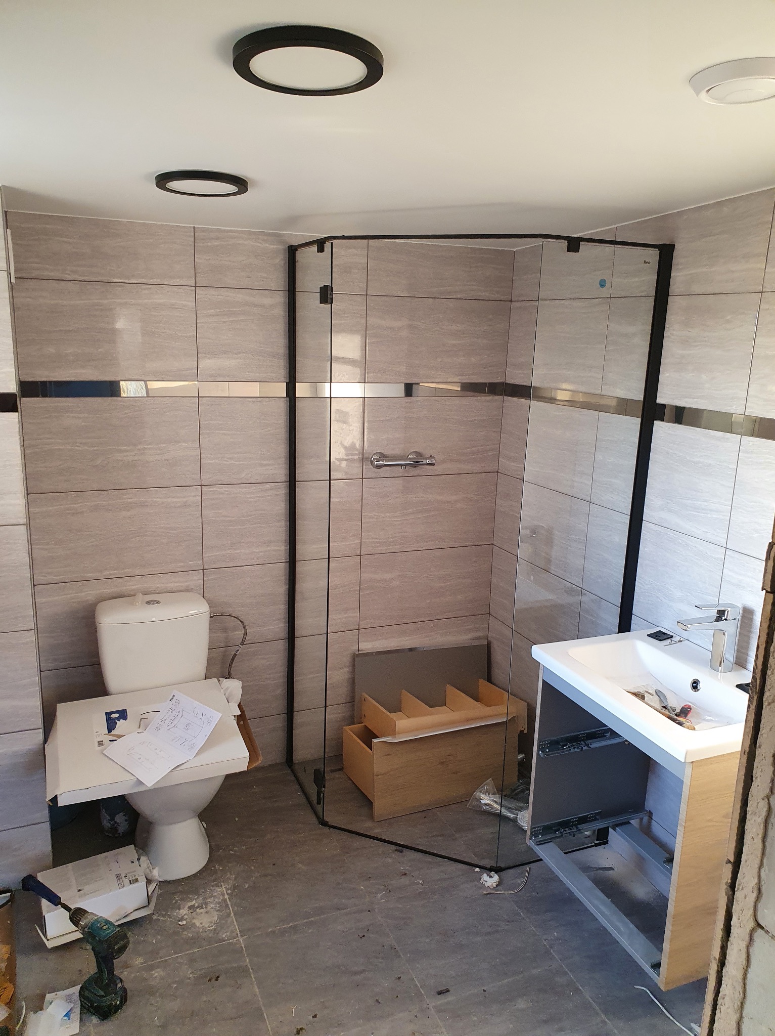 Remont łazienki: montaż kabiny prysznicowej z czarnymi profilami i umywalki z szafką w trakcie instalacji, widoczne narzędzia i materiały budowlane, płytki imitujące kamień na ścianach.