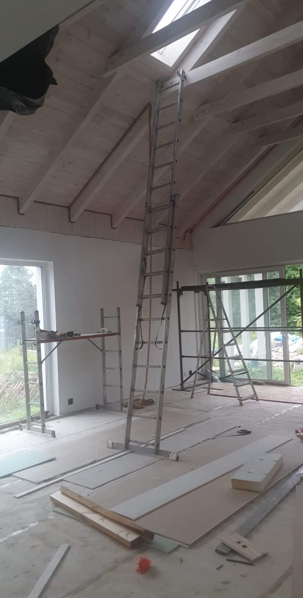 Wnętrze poddasza w trakcie prac: widoczne drewniane belki stropowe, okno dachowe, drabina oparta o sufit, rusztowania oraz płyty gipsowo-kartonowe na podłodze.