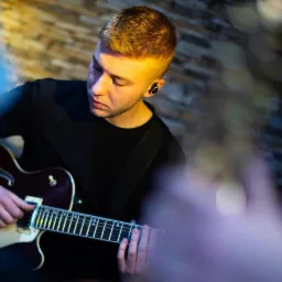 Młody gitarzysta w czarnym T-shircie gra na gitarze elektrycznej o ciemnej barwie, z słuchawkami dousznymi, na tle ściany z cegły.