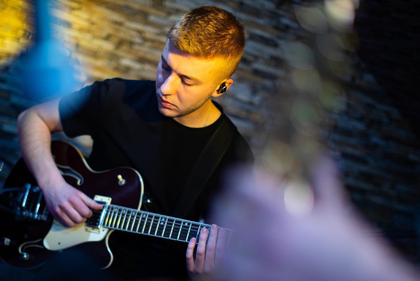 Młody gitarzysta w czarnym T-shircie gra na gitarze elektrycznej o ciemnej barwie, z słuchawkami dousznymi, na tle ściany z cegły.