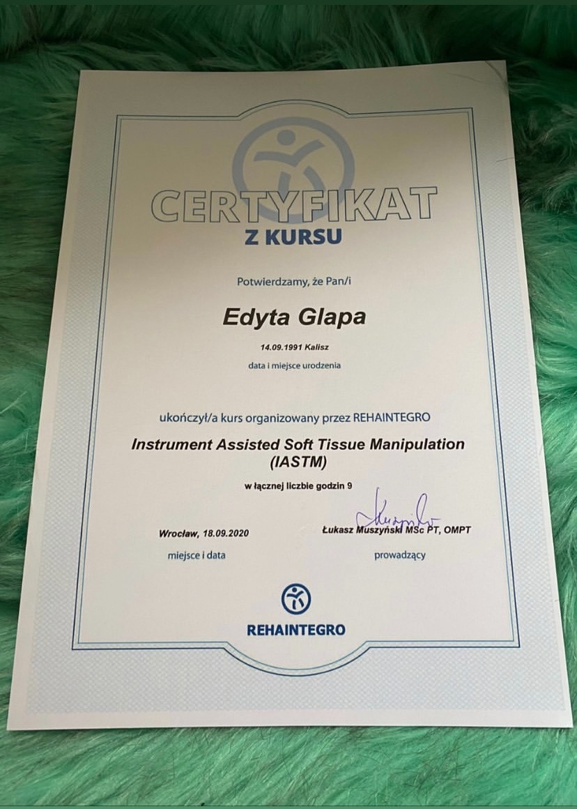 Certyfikat ukończenia kursu Instrument Assisted Soft Tissue Manipulation (IASTM) przez Edytę Glapę, wydany przez REHAINTEGRO z podpisem Łukasza Muszyńskiego, na tle zielonego futerka.