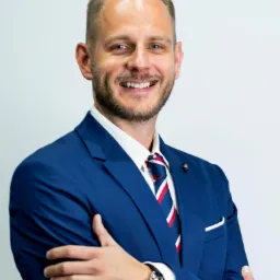 Uśmiechnięty mężczyzna w niebieskim garniturze i krawacie w biało-czerwone paski, z zegarkiem na ręce, pozujący na jasnym tle.