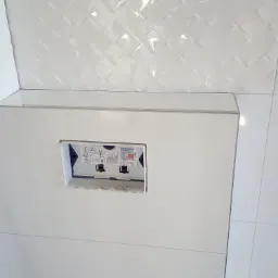 Biała łazienka w trakcie prac wykończeniowych, widoczna zabudowa stelaża podtynkowego WC z otworem na przycisk spłukujący i otwory na rury, błyszczące płytki na suficie ułożone w romby.