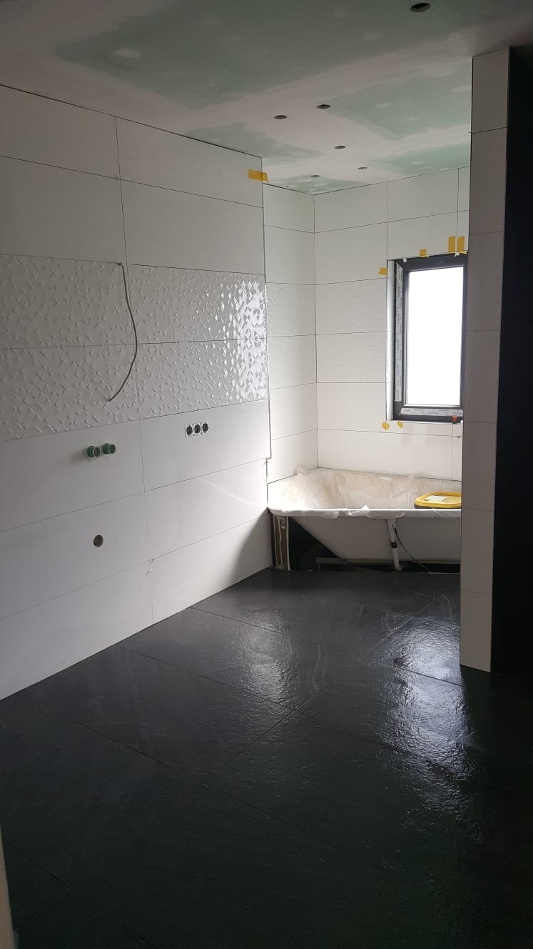 Łazienka w trakcie prac: białe płytki ścienne z dekoracyjnym wzorem, ciemna, błyszcząca podłoga, wanna zabezpieczona folią, widoczne przygotowane punkty instalacji.