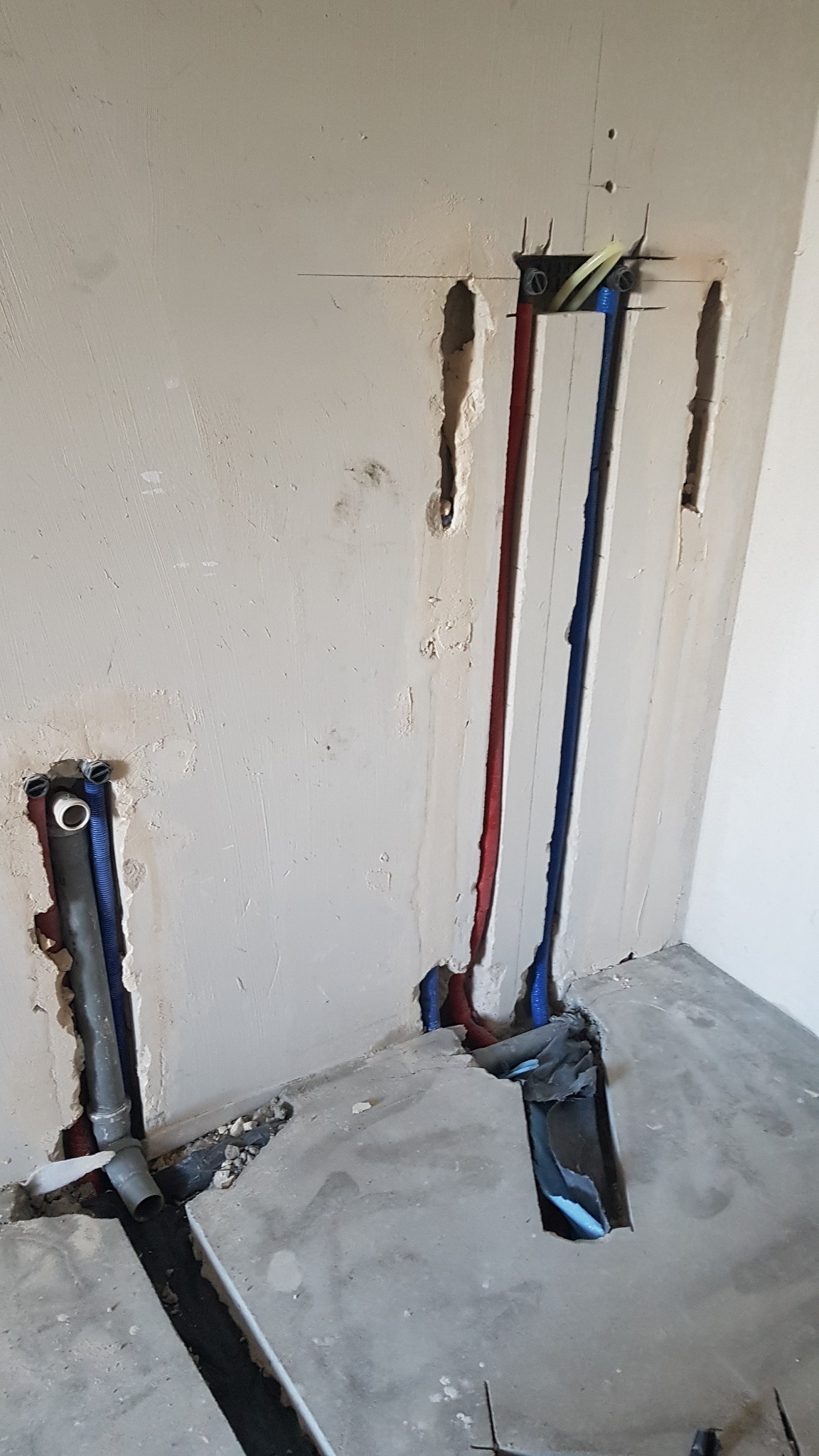 Świeżo wycięte bruzdy w ścianie i posadzce pod pionowe rury wodne (czerwona i niebieska) oraz szarą rurę kanalizacyjną, widoczne ślady po wierceniu i kuciu w betonie i tynku.