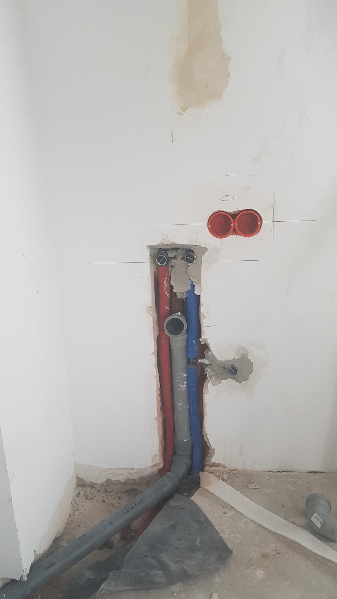 Wnęka w ścianie z widocznymi rurami instalacji wodnej (czerwona i niebieska) oraz kanalizacyjnej (szara) w trakcie prac montażowych, z widocznymi śladami po kuciu i zaprawie.