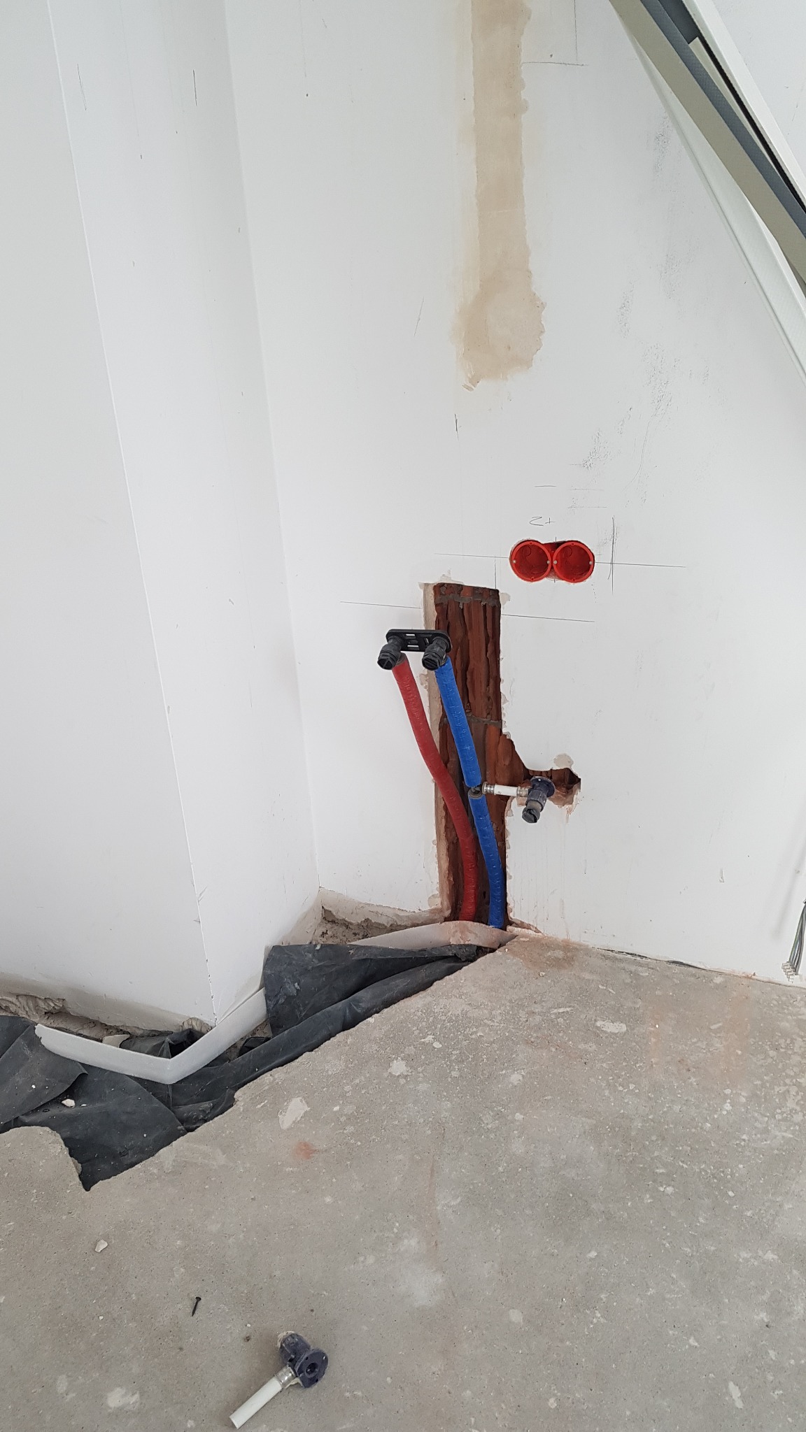 Wnęka w ścianie z wyprowadzonymi rurami instalacji wodnej (czerwona i niebieska) oraz elementami mocującymi, widoczne ślady po kuciu w ścianie, fragment podłogi z czarną folią ochronną i białą rurką.