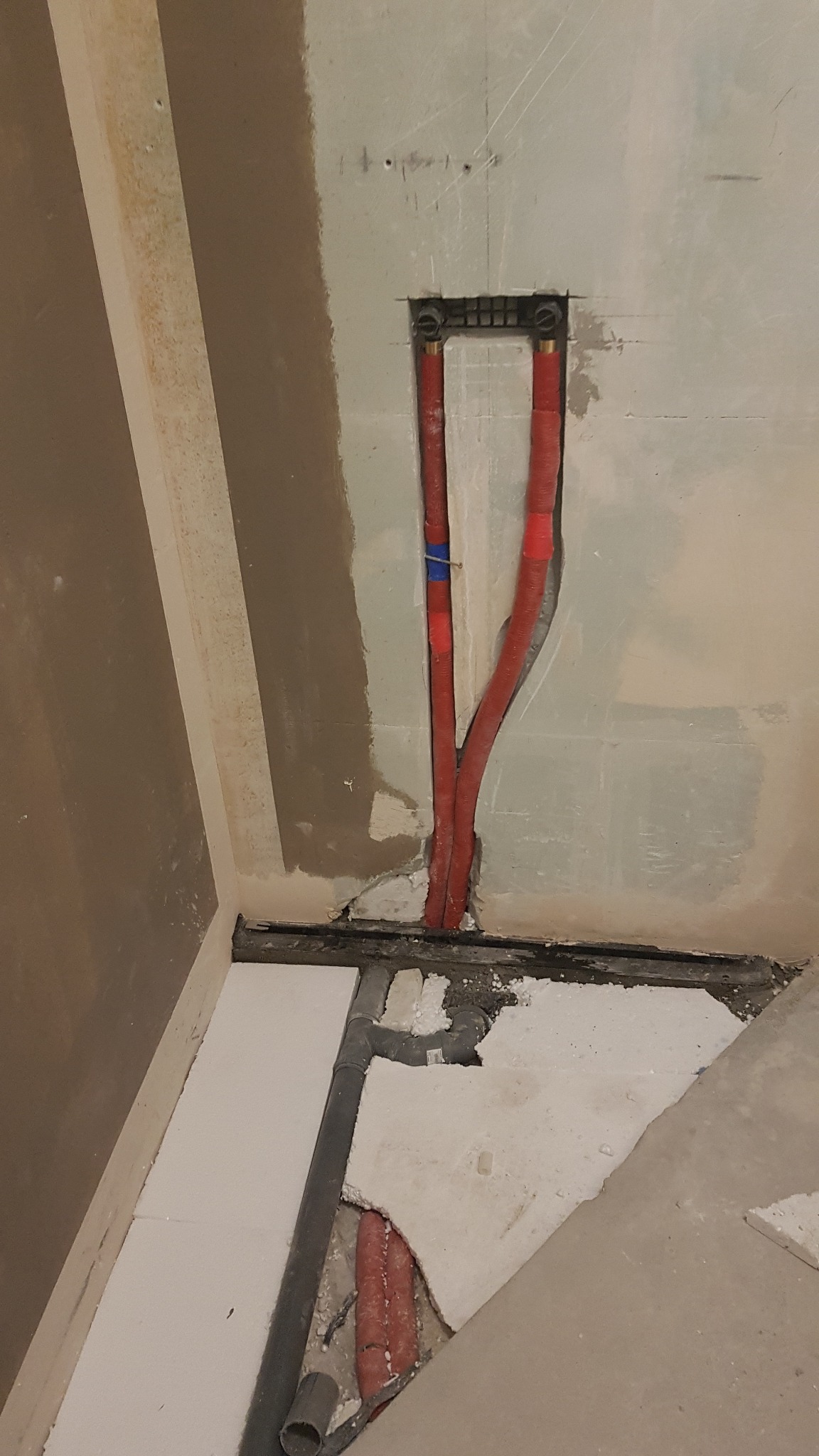 Fragment ściany w trakcie remontu z widocznymi czerwonymi rurami instalacji wodnej wychodzącymi z puszki podtynkowej, szara rura odpływowa w podłodze i fragmenty styropianu.