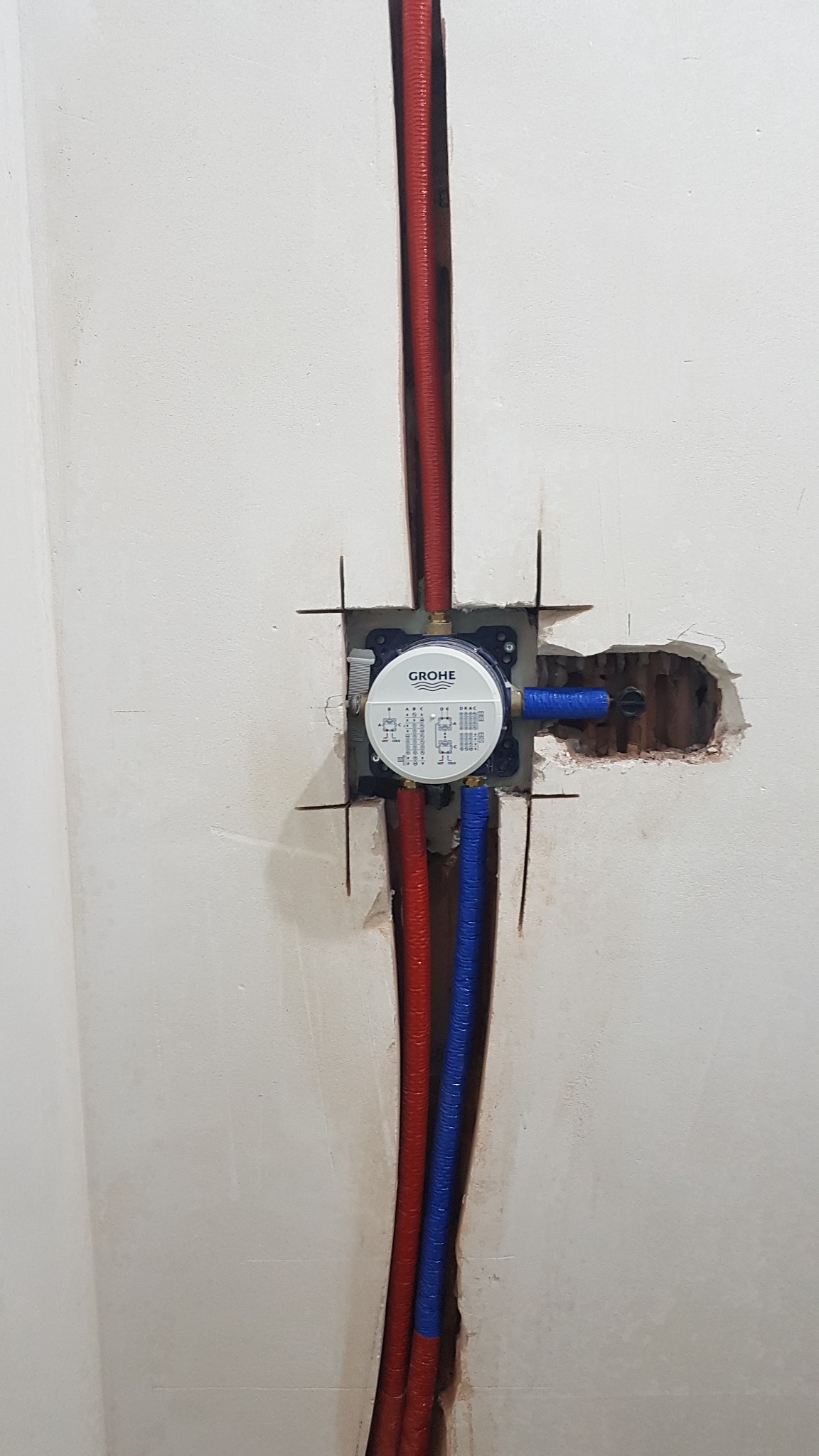 Ściana z zamontowanym podtynkowym zaworem Grohe, z widocznymi rurami doprowadzającymi wodę w kolorze czerwonym i niebieskim, otoczenie w stanie surowym przed wykończeniem.