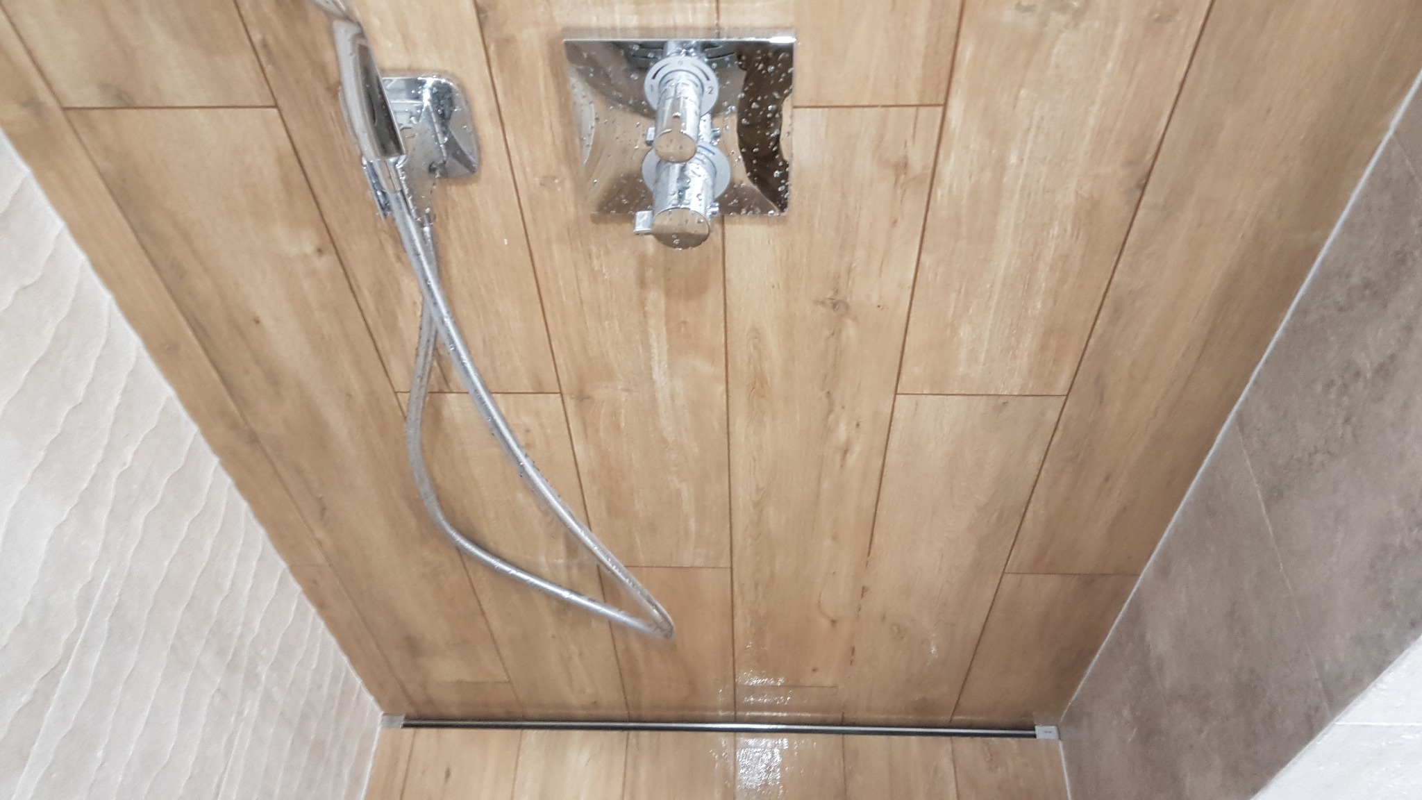 Nowoczesna kabina prysznicowa z odpływem liniowym, wykończona płytkami imitującymi drewno, chromowana armatura i słuchawka prysznicowa.