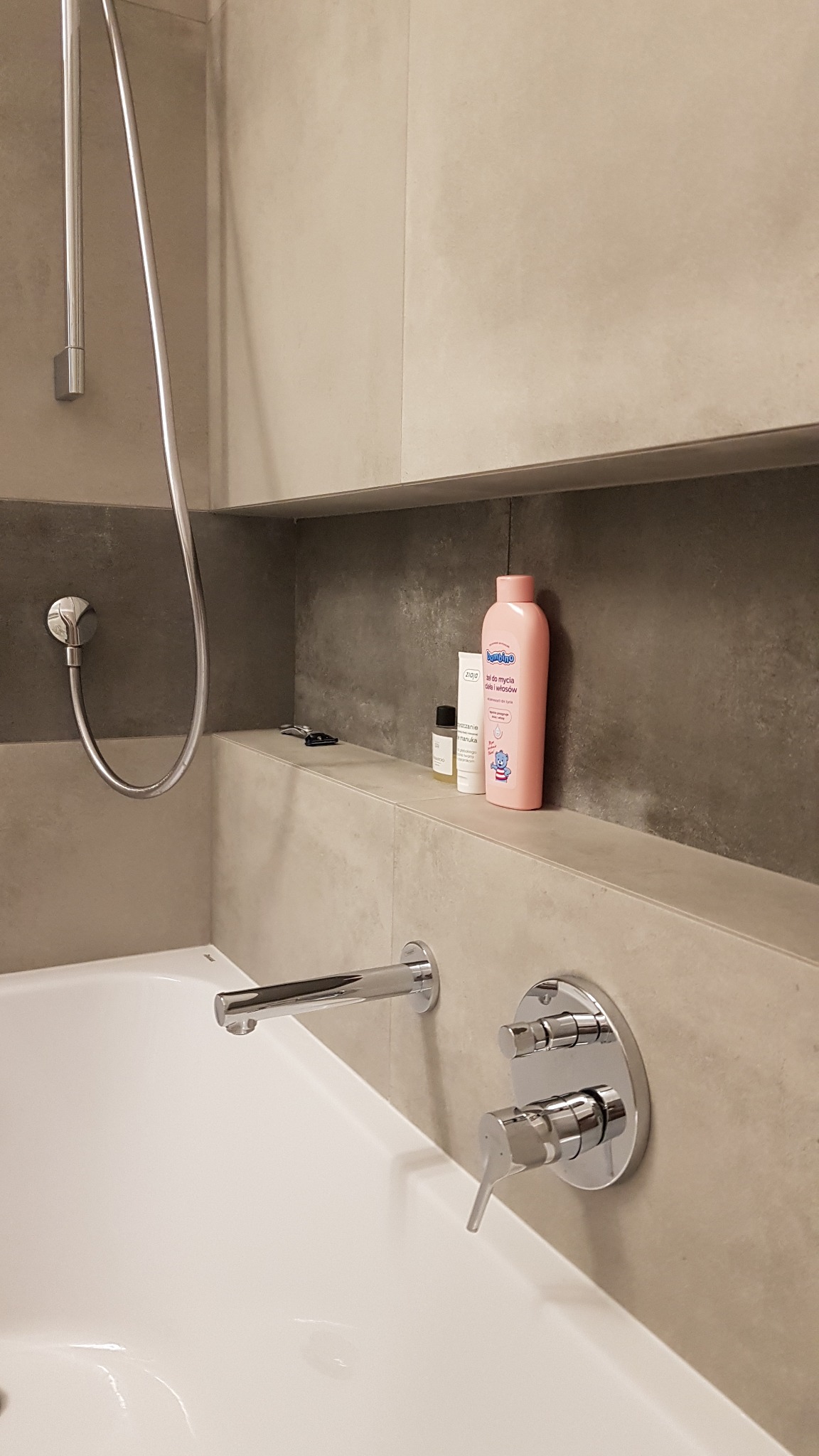 Nowoczesna bateria wannowa chromowana, zamontowana na beżowej ścianie z półką, na której stoją kosmetyki; widoczna słuchawka prysznicowa.