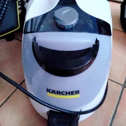 Parownica Kärcher w kolorze białym i szarym, stojąca na terakotowej podłodze, z widocznym przewodem zasilającym.