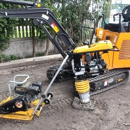 BOGMAR - Pomarańczowa mini-koparka z łyżką, żółta zagęszczarka płytowa i stopowa na placu budowy, obok betonowego płotu i zieleni.