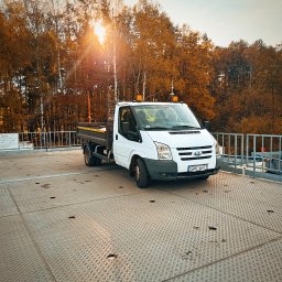 BOGMAR - Biały Ford Transit z otwartą skrzynią ładunkową i pomarańczowymi lampami ostrzegawczymi, stojący na metalowej nawierzchni, oświetlony zachodzącym słońcem przez jesienne drzewa.