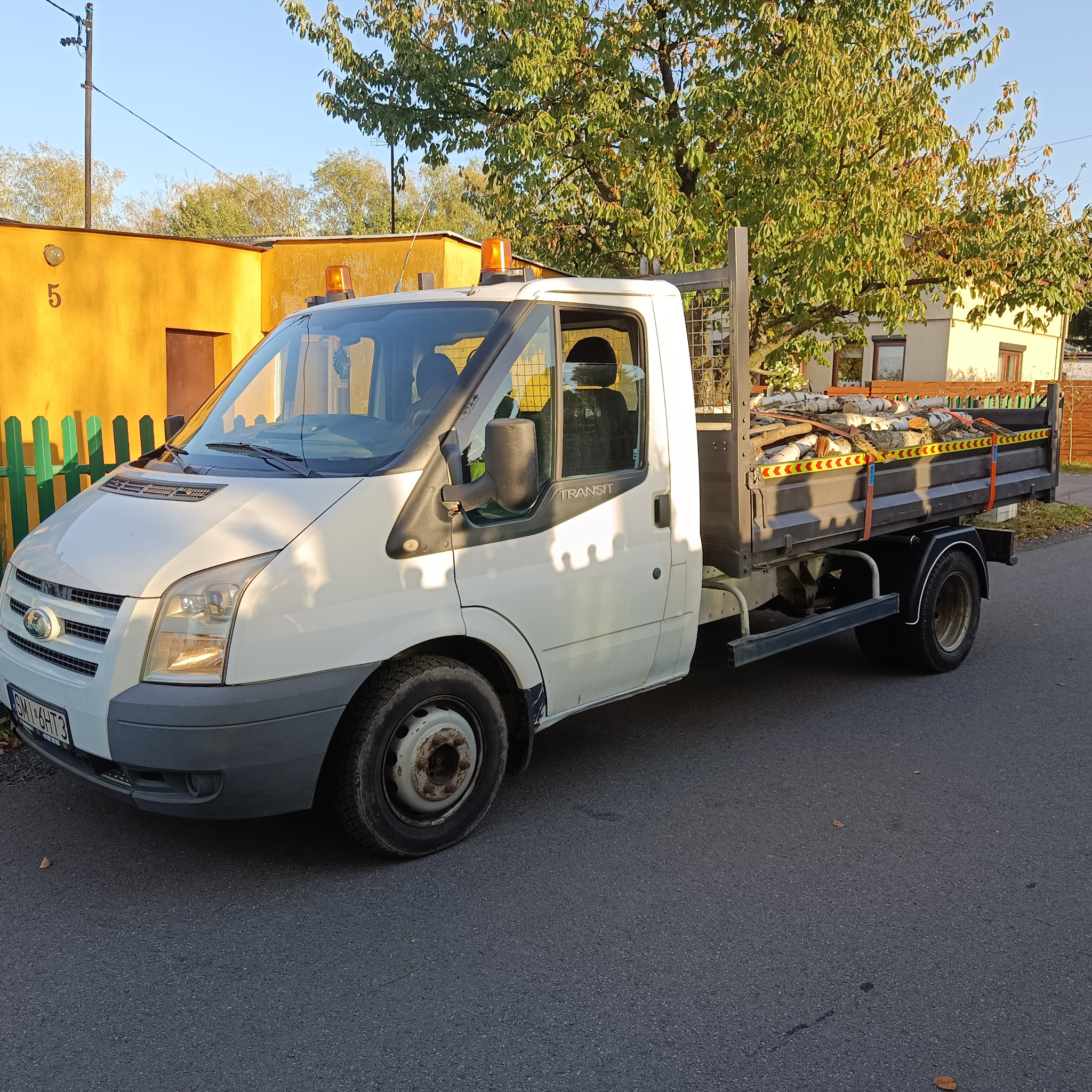 Biały samochód dostawczy Ford Transit z pomarańczowym światłem ostrzegawczym, zaparkowany na asfaltowej drodze, z ładunkiem pociętych pni drzew zabezpieczonych pasami w skrzyni ładunkowej