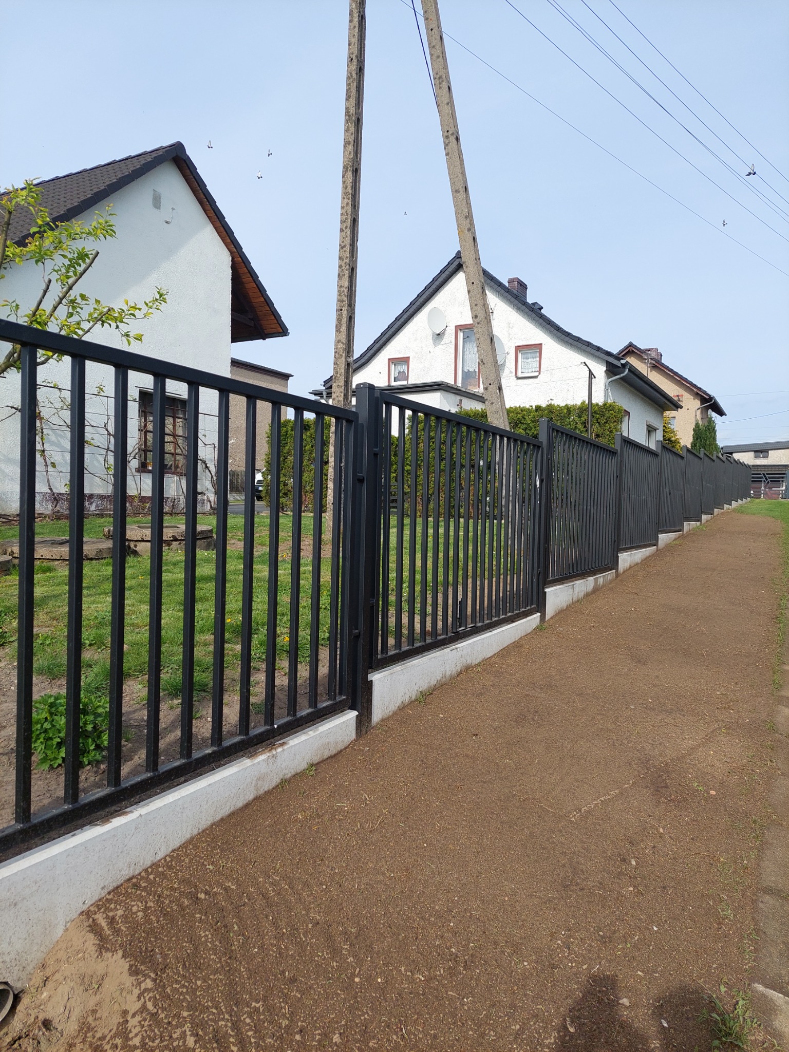 Długa, czarna, metalowa ogrodzenie panelowe z betonową podmurówką, oddzielające posesję od przygotowanego podłoża pod trawnik, z widocznymi domami w tle.