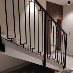 Nowoczesna klatka schodowa z biało-szarymi schodami i czarną, geometryczną balustradą. Widoczne zabezpieczenia na stopniach i elementy wykończenia wnętrza.