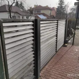 Metalowa brama wjazdowa lamelowa, widok z bliska na szarą konstrukcję z poziomych listew na tle chodnika z kostki, ulicy i zabudowań.