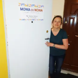 Kobieta, prawdopodobnie właścicielka, stoi przy otwartych drzwiach gabinetu logopedycznego z napisem 'Mowa od Nowa' i danymi kontaktowymi.