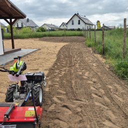 MUCHA NIE SIADA - USŁUGI OGRODNICZE ALICJA MUCHA - Przygotowanie terenu pod trawnik: glebogryzarka spulchnia piaszczystą ziemię wzdłuż ogrodzenia, widoczne ślady opon i budynki w tle.