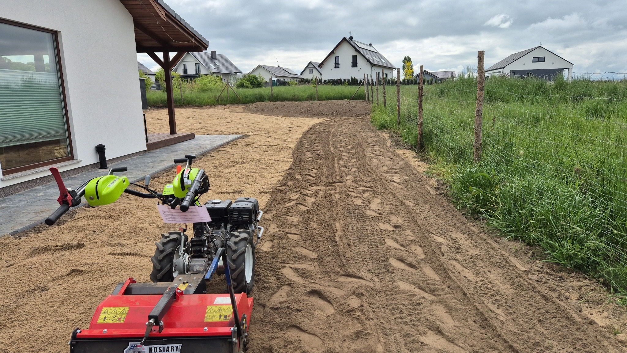 Przygotowanie terenu pod trawnik: glebogryzarka spulchnia piaszczystą ziemię wzdłuż ogrodzenia, widoczne ślady opon i budynki w tle.