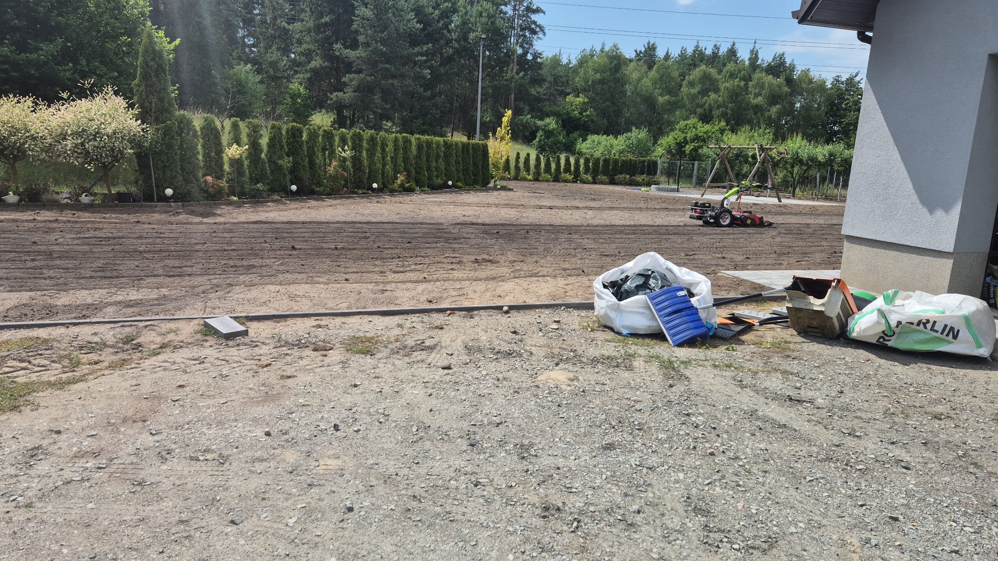 Przygotowany teren pod trawnik z widoczną glebogryzarką, workami na ziemię i narzędziami ogrodniczymi. W tle zadbany żywopłot i drzewa.