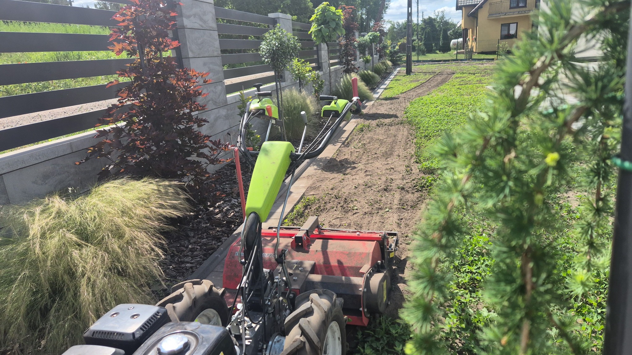 Przygotowanie gleby pod trawnik: glebogryzarka spalinowa na działce, widok z perspektywy operatora, świeżo przekopana ziemia, obok rosną drzewa i krzewy.