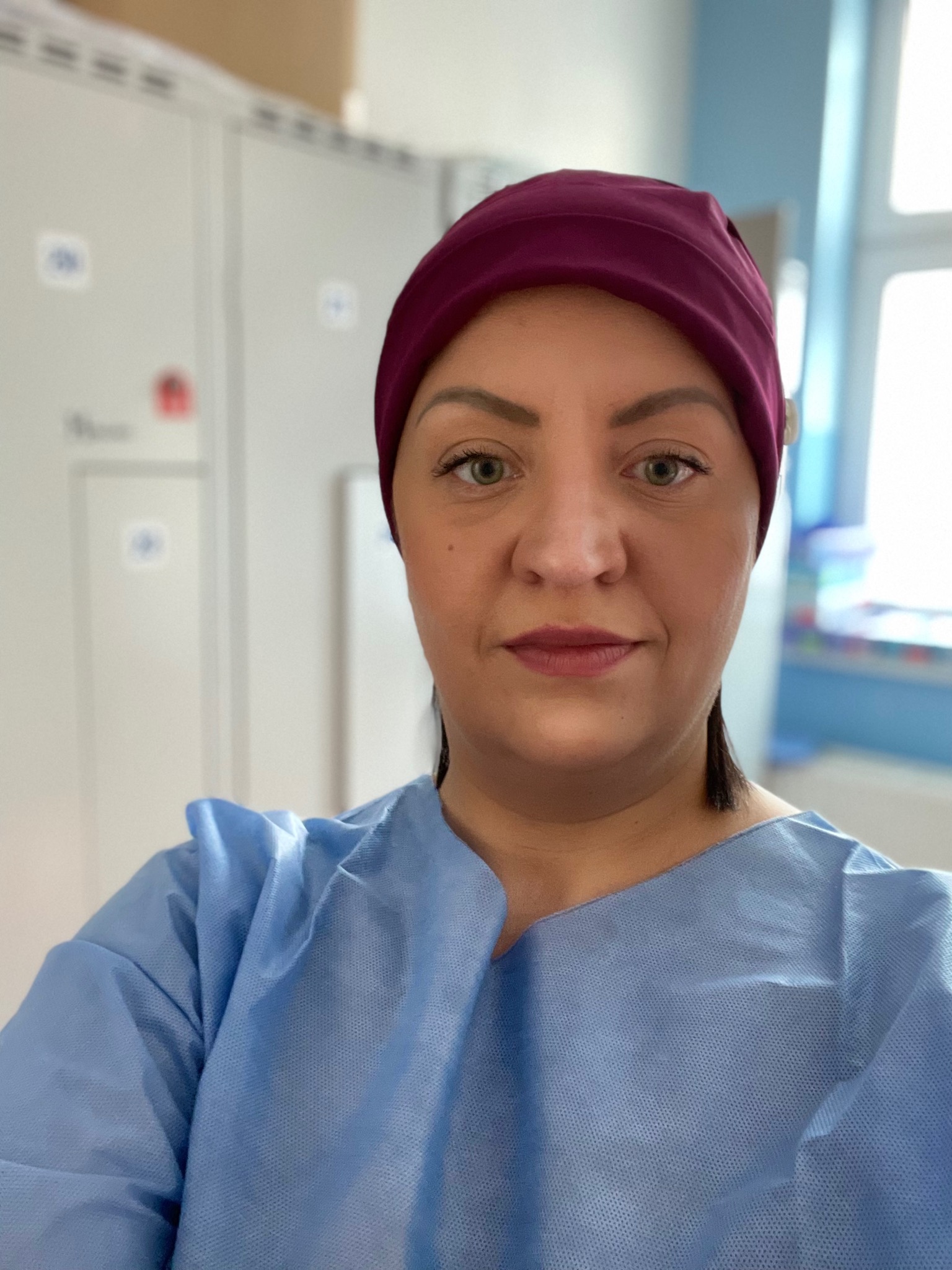 Portret pielęgniarki w niebieskim uniformie i bordowym czepku medycznym, z zielonymi oczami, na tle białych szafek i okna.