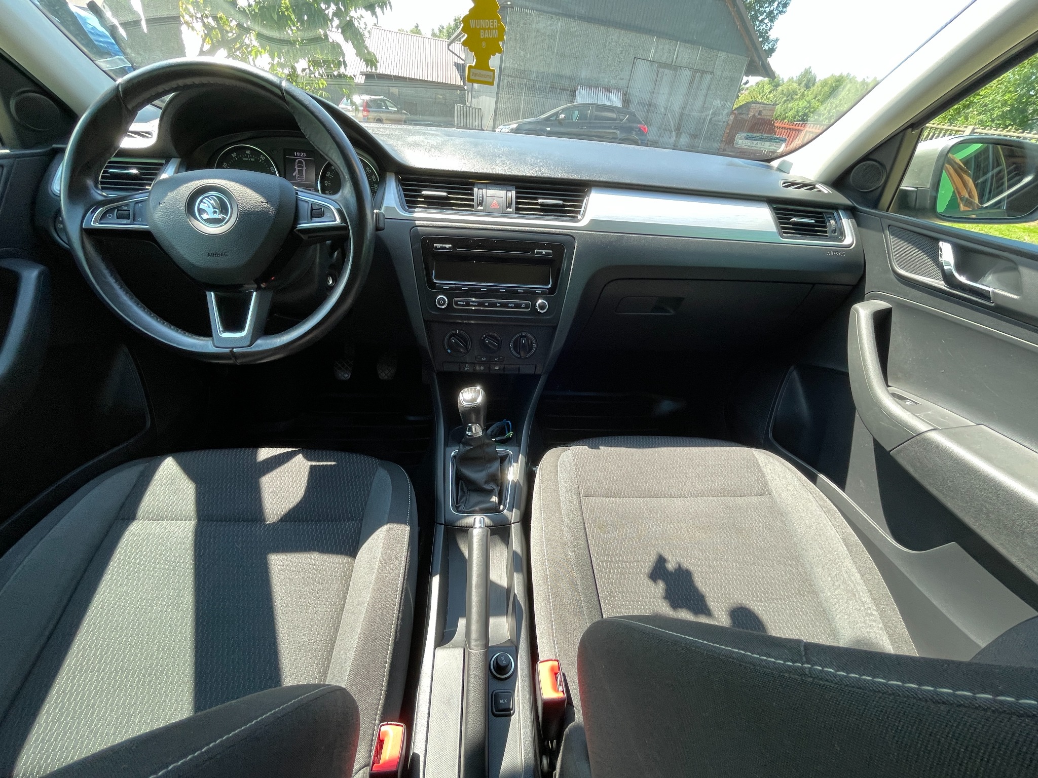 Wnętrze samochodu marki Skoda Rapid z widocznym kokpitem, kierownicą z logo, szarą tapicerką foteli oraz zawieszką zapachową Wunder-Baum na lusterku.