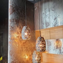 Geo Glazura profesjonalne glazurnictwo - Trzy geometryczne lampy wiszące z podświetleniem na tle ściany z efektem rdzy i drewnianej półki. Nowoczesny design oświetlenia wnętrza.