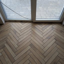 Geo Glazura profesjonalne glazurnictwo - Ułożona w jodełkę podłoga z płytek imitujących drewno, widok z góry, z fragmentem okna balkonowego i kostką brukową na zewnątrz. Częściowo ukończona realizacja.
