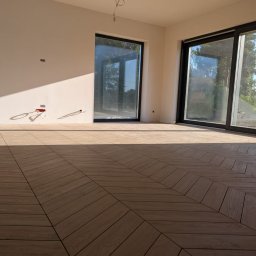 Geo Glazura profesjonalne glazurnictwo - Jasne płytki podłogowe ułożone we wzór jodełki w przestronnym, słonecznym wnętrzu z dużymi oknami. Widoczne niedokończone prace przy krawędzi podłogi.