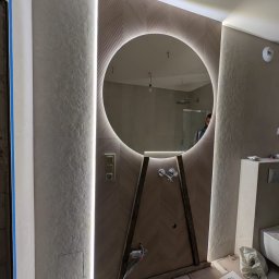 Geo Glazura profesjonalne glazurnictwo - Nowoczesna łazienka w trakcie wykańczania: okrągłe lustro z podświetleniem LED, wzorzysta ściana, widoczne elementy instalacji wodnej i elektrycznej, surowy stan podłogi.