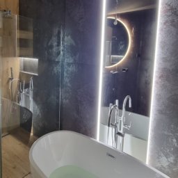 Geo Glazura profesjonalne glazurnictwo - Nowoczesna łazienka z wolnostojącą wanną wypełnioną wodą, podświetlanym lustrem w kształcie półksiężyca i szklaną kabiną prysznicową. Ciemne płytki i drewniane akcenty.