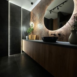 Geo Glazura profesjonalne glazurnictwo - Nowoczesna łazienka z podświetlanym lustrem owalnym, drewnianą szafką pod umywalką i ciemnymi płytkami na ścianach oraz podłodze. Minimalistyczny design z roślinami.