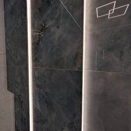 Geo Glazura profesjonalne glazurnictwo - Nowoczesne wykończenie ściany z ciemnych, wzorzystych płytek imitujących onyks, z wbudowanym pionowym oświetleniem LED i subtelnymi geometrycznymi akcentami świetlnymi.