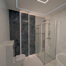 Geo Glazura profesjonalne glazurnictwo - Nowoczesna łazienka z białymi płytkami marmurowymi, ciemną ścianą akcentową z pionowymi listwami LED, szklaną kabiną prysznicową, podwieszaną toaletą i wanną wolnostojącą. Sufit z geometrycznym...