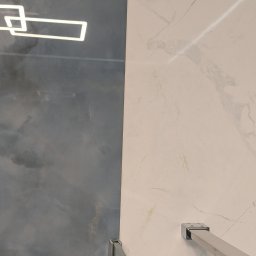 Geo Glazura profesjonalne glazurnictwo - Fragment nowoczesnej łazienki z ciemną, teksturowaną ścianą i jasnymi płytkami imitującymi marmur, widoczne chromowane mocowanie kabiny prysznicowej oraz odbicie prostokątnej lampy.