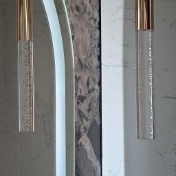 Geo Glazura profesjonalne glazurnictwo - Dwie nowoczesne lampy wiszące ze złotymi elementami i cylindrycznymi kloszami z bąbelkami, obok podświetlanego lustra i marmurowych ścian. Eleganckie wykończenie wnętrza.