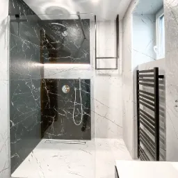 Nowoczesna łazienka z kabiną prysznicową typu walk-in wyłożoną czarnym marmurem z białymi żyłami, białe marmurowe ściany i podłoga, czarny grzejnik drabinkowy i wieszak na ręczniki.