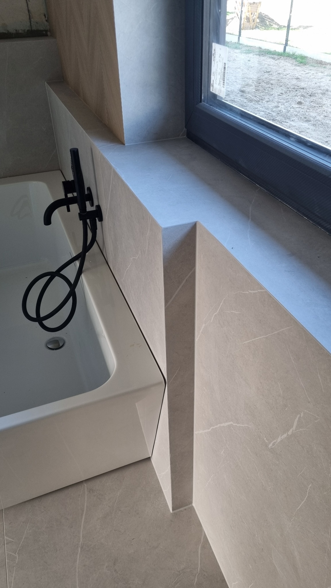 Wykończona łazienka z wanną, czarną armaturą i oknem. Płytki imitujące marmur na ścianach i podłodze. Geometryczne wykończenie wnęki okiennej.