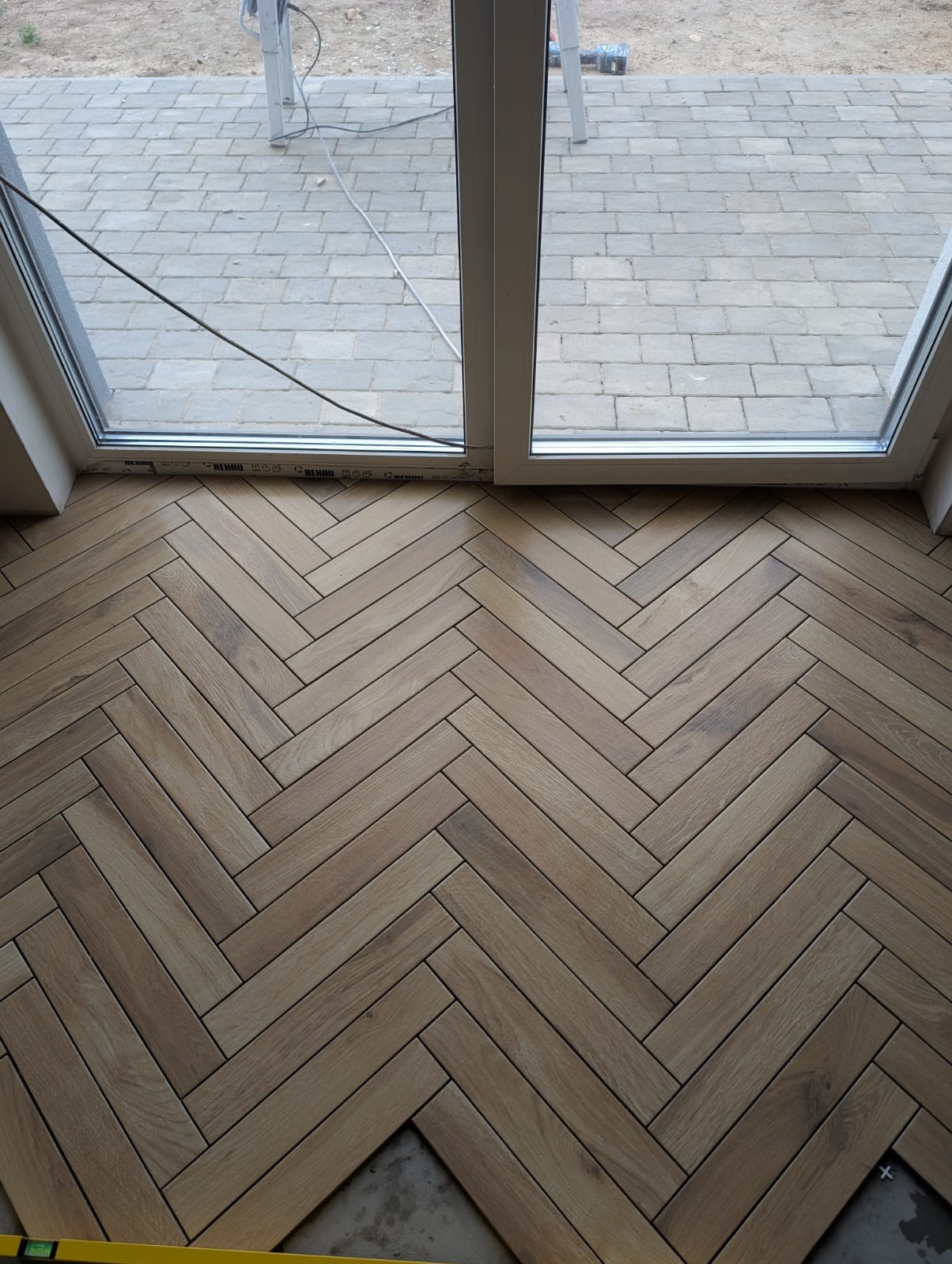 Ułożona w jodełkę podłoga z płytek imitujących drewno, widok z góry, z fragmentem okna balkonowego i kostką brukową na zewnątrz. Częściowo ukończona realizacja.