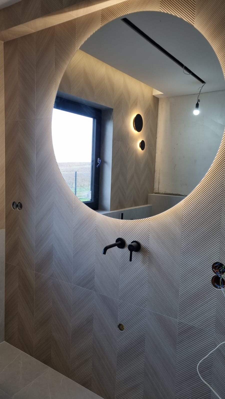Łazienka z okrągłym lustrem z podświetleniem LED i wzorem jodełki na płytkach. Widoczne czarne baterie i okno z widokiem na zewnątrz. Częściowo wykończone wnętrze.