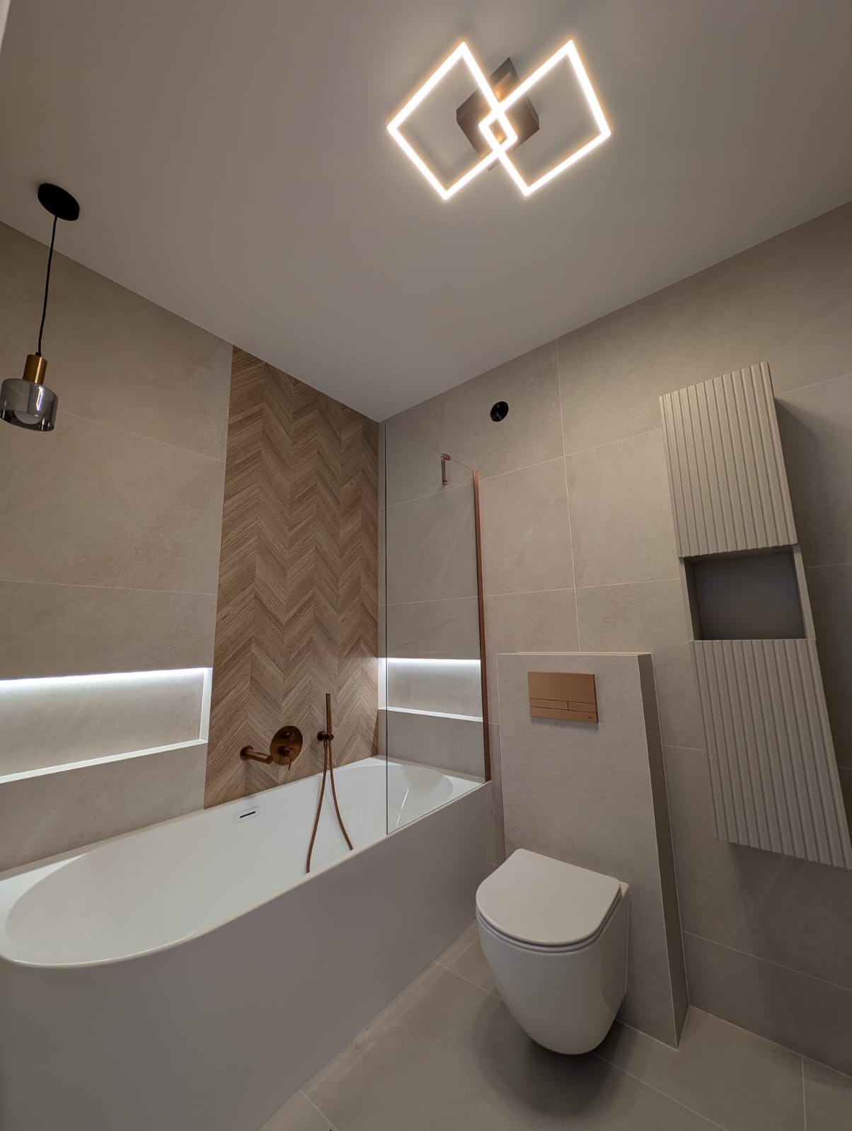 Nowoczesna łazienka z wanną, toaletą i grzejnikiem dekoracyjnym. Uwagę przykuwają płytki ułożone w jodełkę oraz oświetlenie LED. Całość utrzymana w stonowanej kolorystyce.